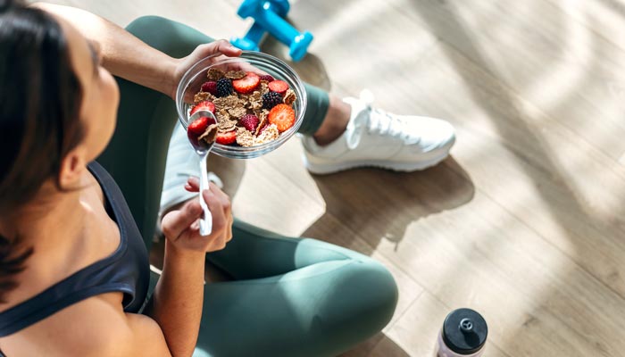 Young woman sitting in gym, eating a bowl of cereal and fruit.