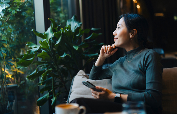 GettyImages-2149615639 young Asian woman with smartphone looking out through window while sitting in a cafe having coffee
